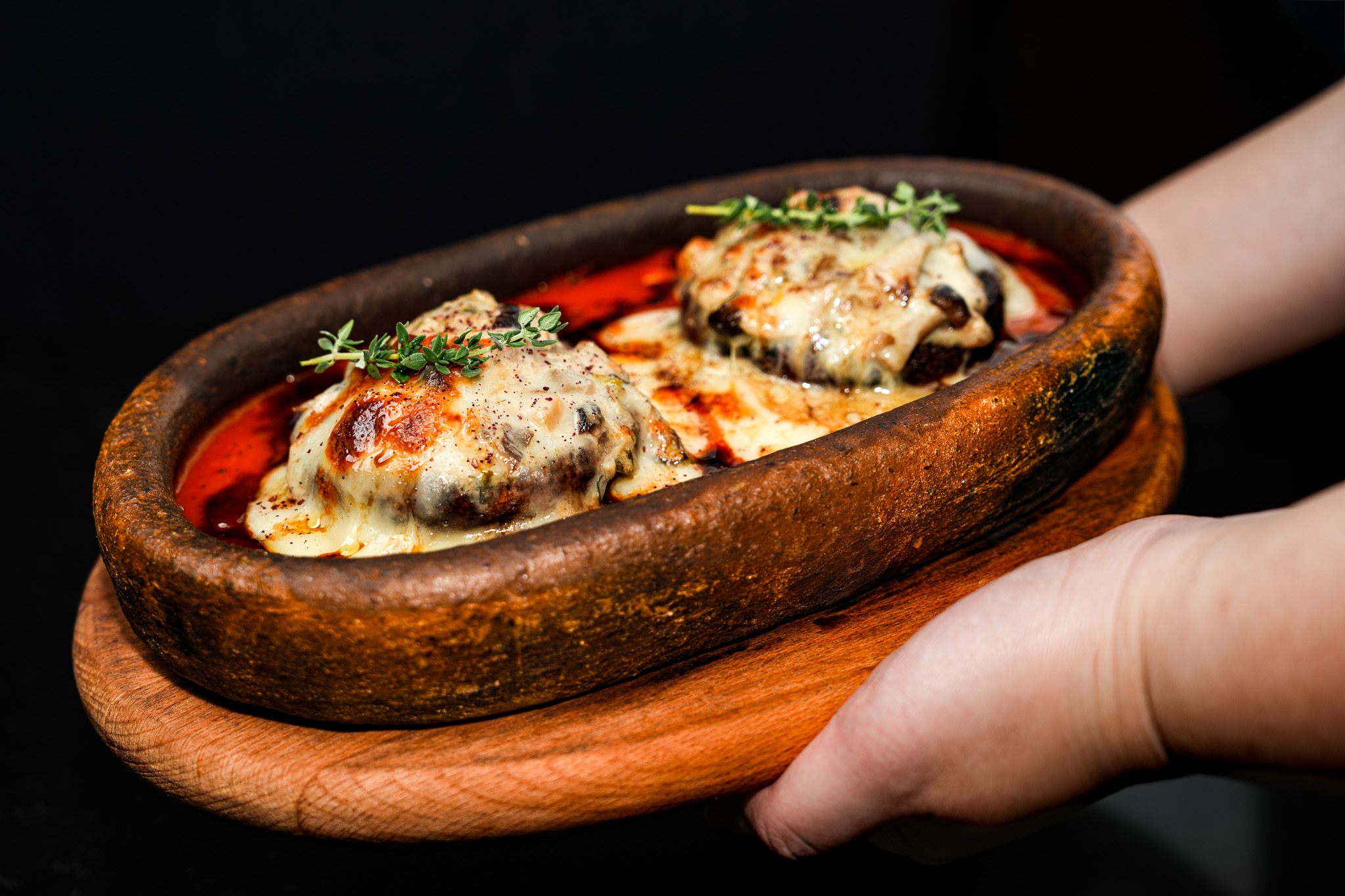 Hands presenting a clay dish of cheese-topped aubergine