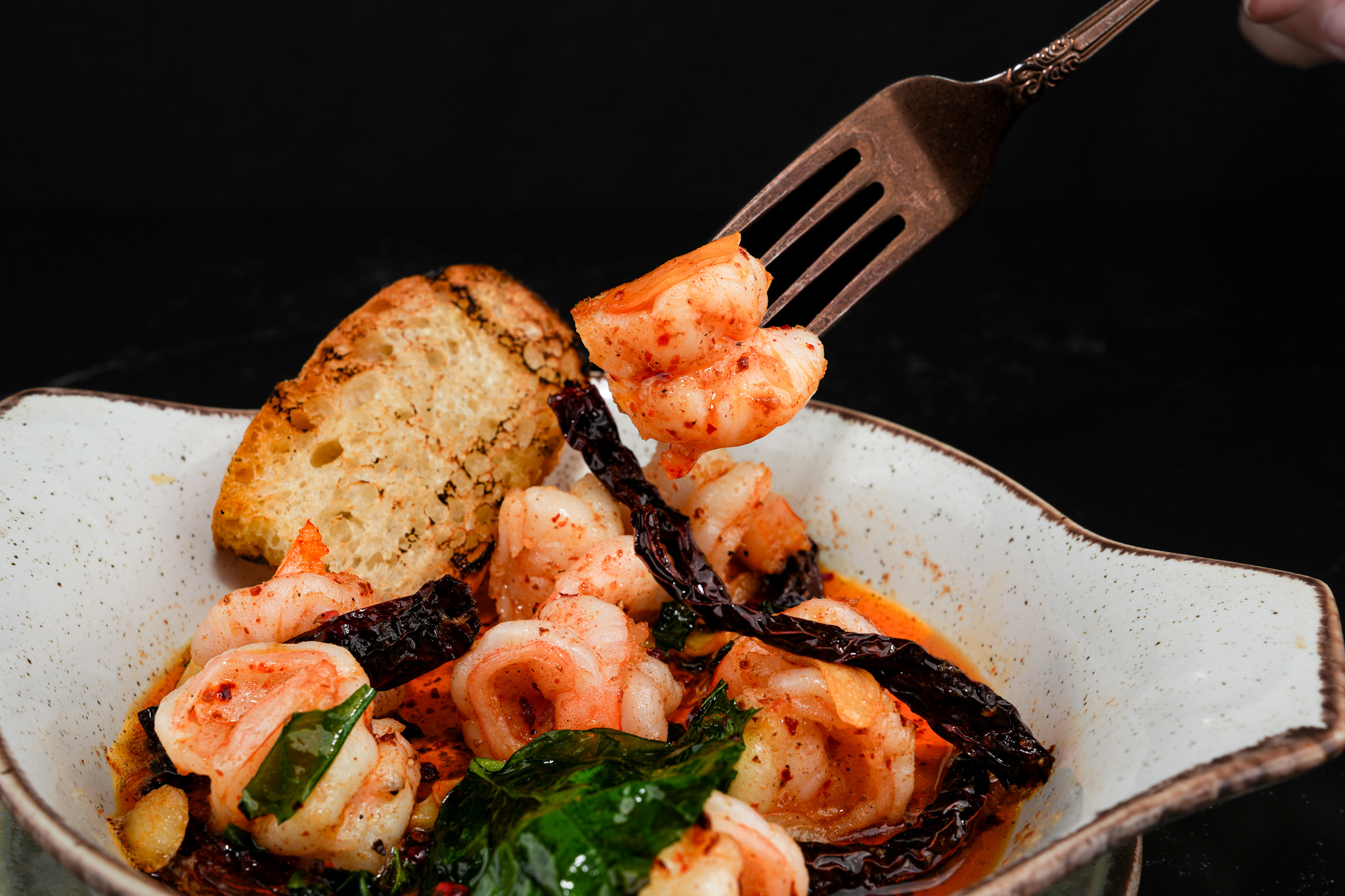 A fork lifting a garlic prawn beside toasted bread