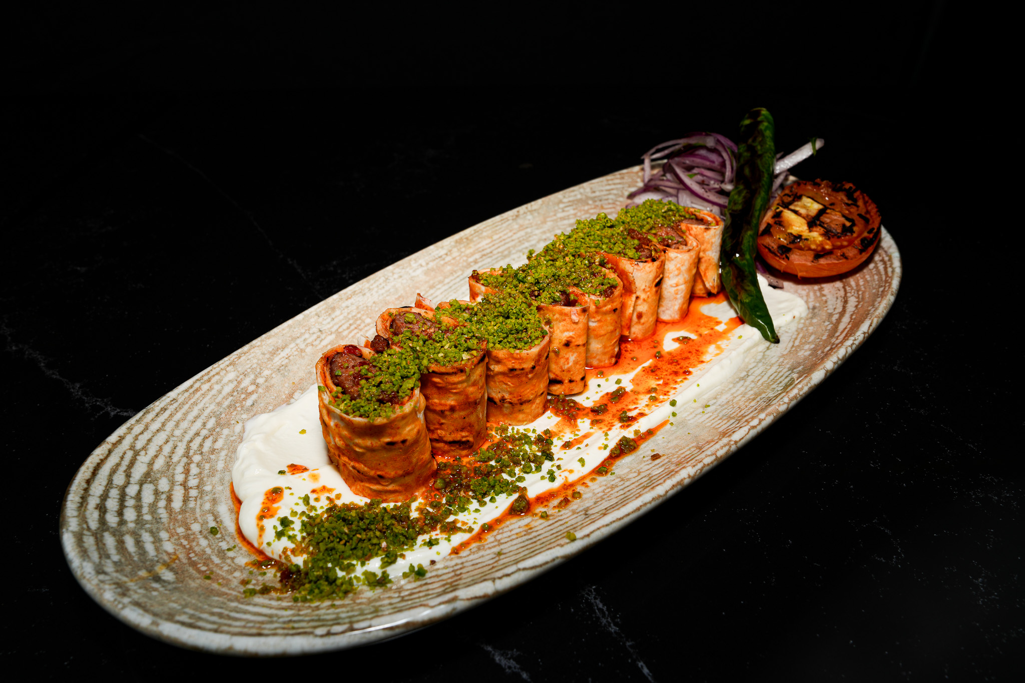 Sliced kebab rolls topped with pistachio over yogurt and sauce