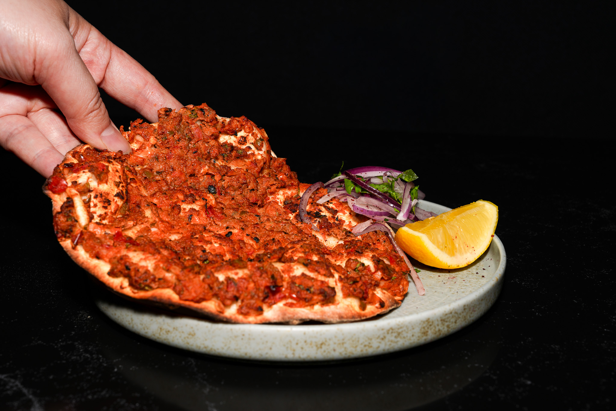A hand lifting lahmacun beside onions and lemon