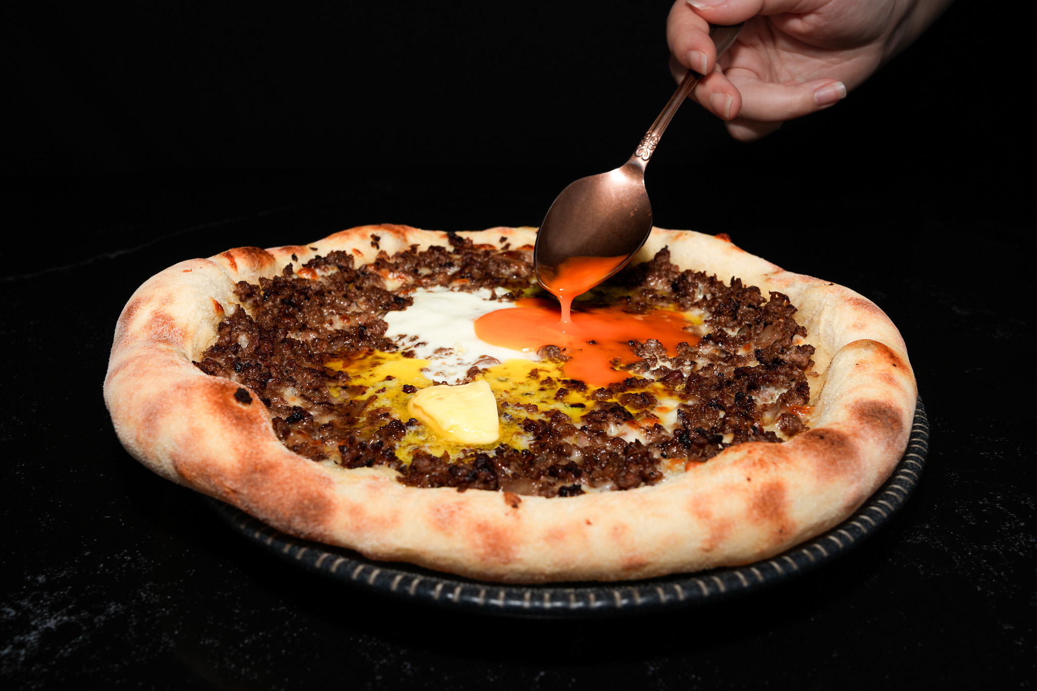 A spoon breaking the egg yolk on a minced meat flatbread
