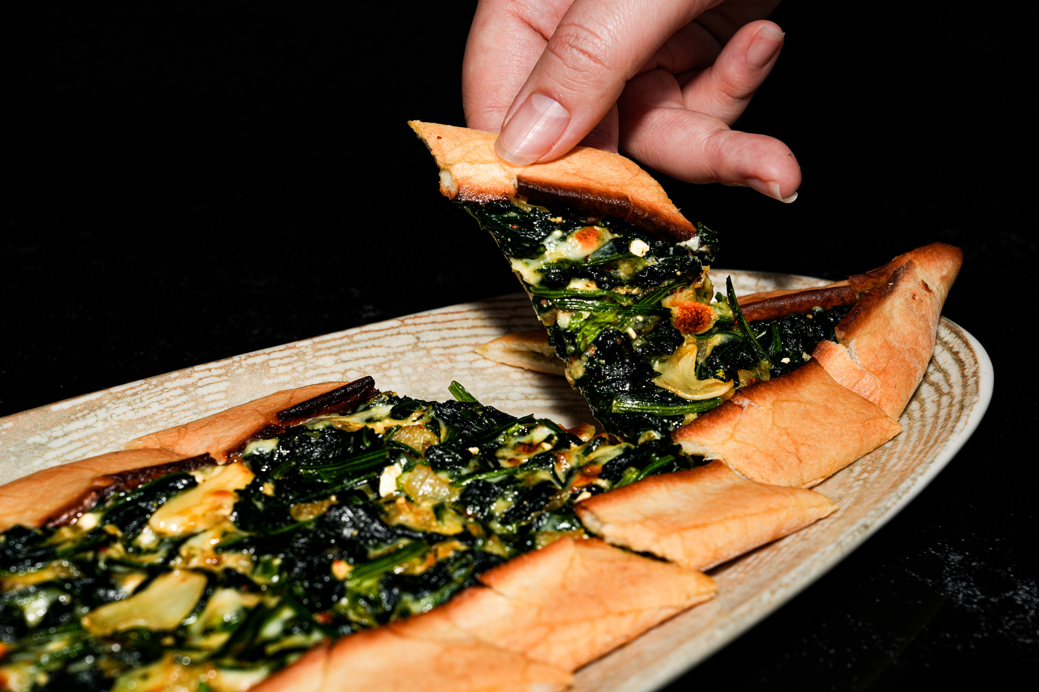 A hand lifting a slice of spinach and cheese pide